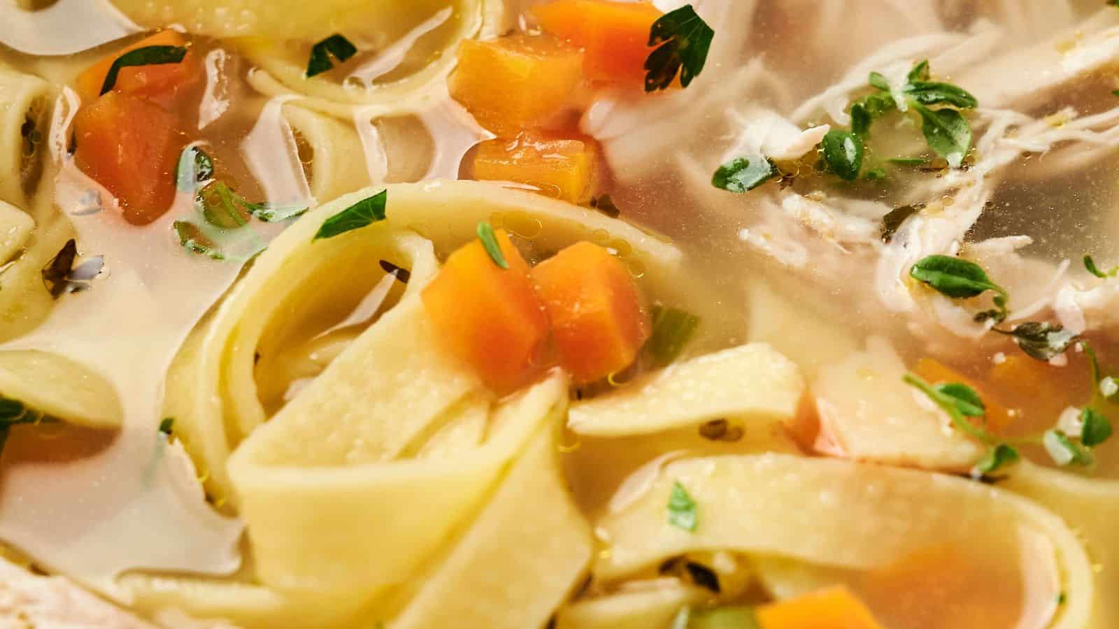 Close-up of chicken noodle soup with wide noodles, diced carrots, pieces of chicken, and fresh herbs in a clear broth.