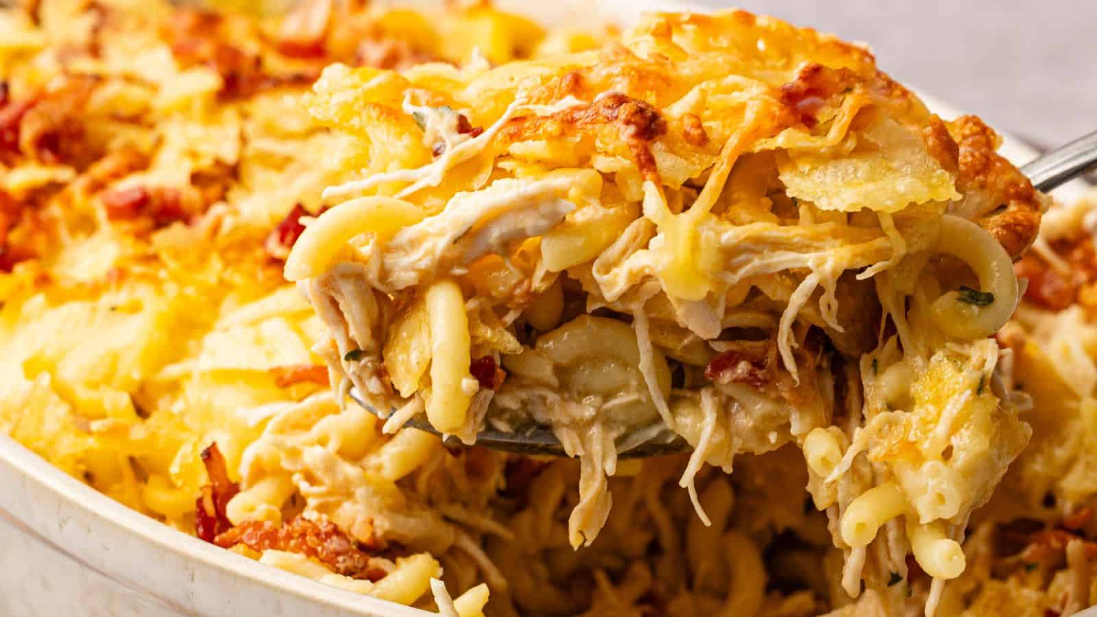 A close-up of a serving spoon lifting cheesy baked pasta with shredded chicken from a casserole dish.