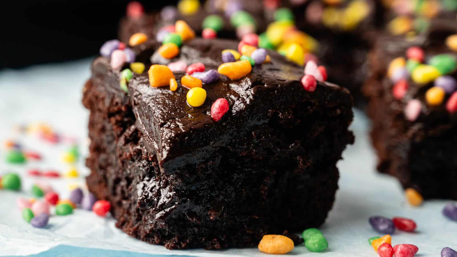 A close-up of a chocolate brownie topped with glossy frosting and colorful candy pieces, resting on parchment paper.