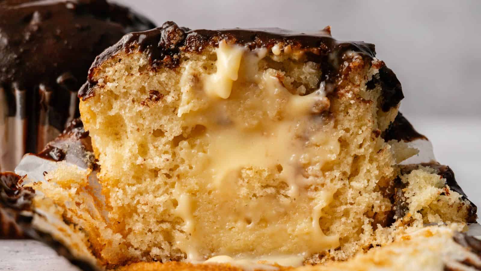 A close-up of a sliced muffin filled with creamy vanilla custard and topped with chocolate glaze.