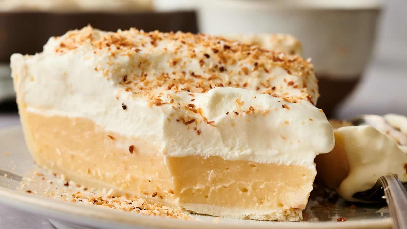 A slice of cream pie with a thick custard filling, topped with whipped cream and toasted coconut flakes, served on a plate.