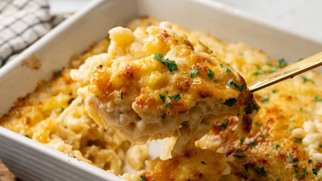 A close-up of a serving of baked macaroni and cheese being lifted from a casserole dish, topped with melted cheese and garnished with chopped herbs.