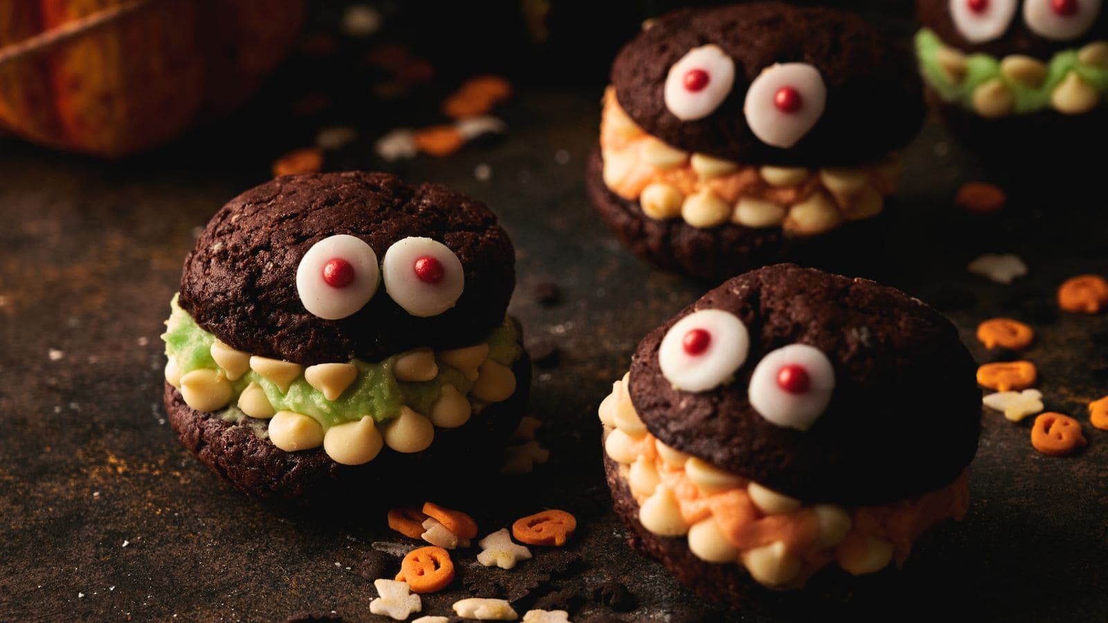 Three chocolate cookie sandwiches decorated with candy eyes and colored frosting to resemble monster faces, surrounded by small Halloween-themed sprinkles.