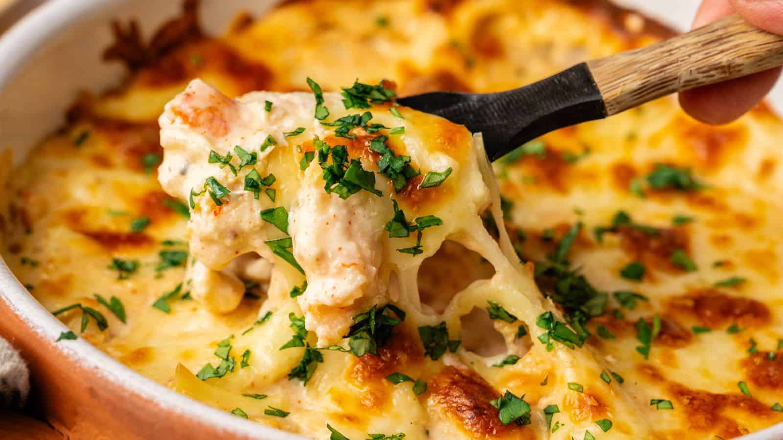 A spoon lifts a portion of cheesy baked pasta topped with chopped parsley from a casserole dish.