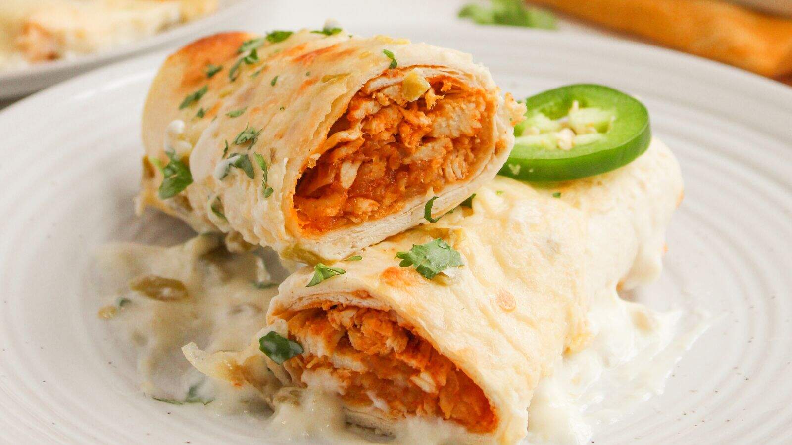 Two tortilla-wrapped enchiladas filled with shredded chicken, topped with creamy white sauce, chopped cilantro, and a jalape&ntilde;o slice, served on a white plate.