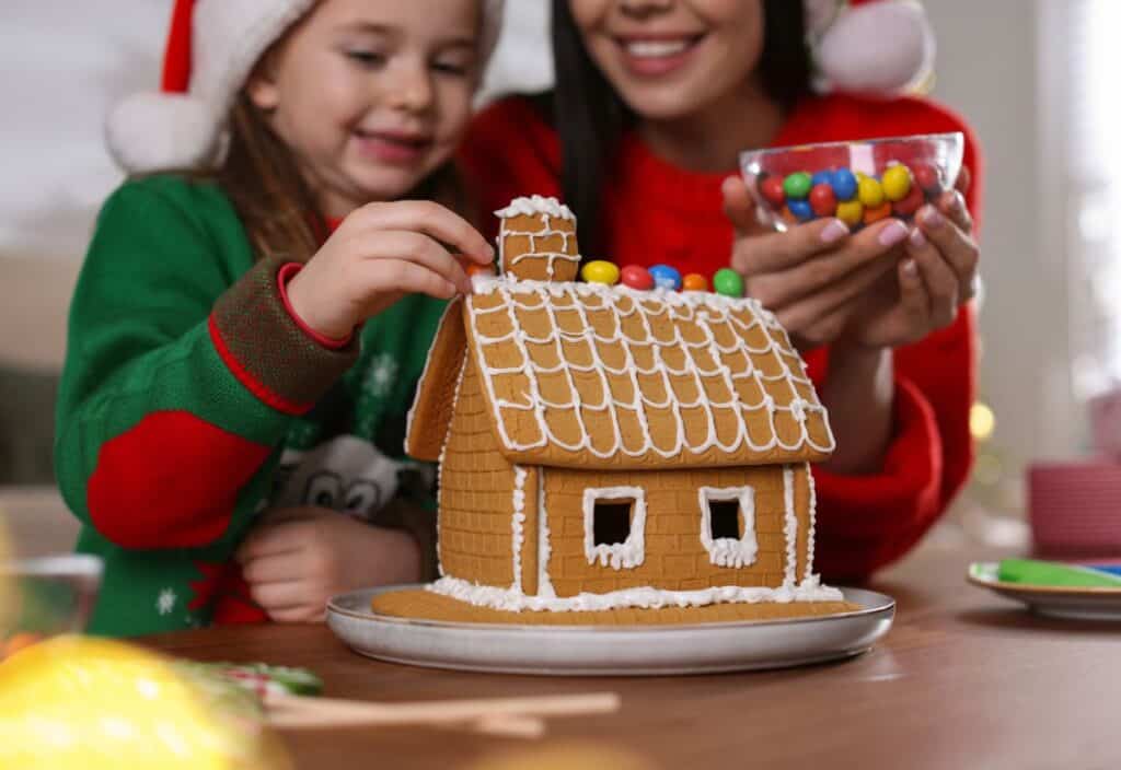 A child and adult decorate a gingerbread house with colorful candies while wearing festive holiday clothing and Santa hats.