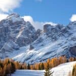 Snow-covered mountain peaks rise above a forest of evergreen and yellow-leaved trees under a blue sky with scattered clouds.