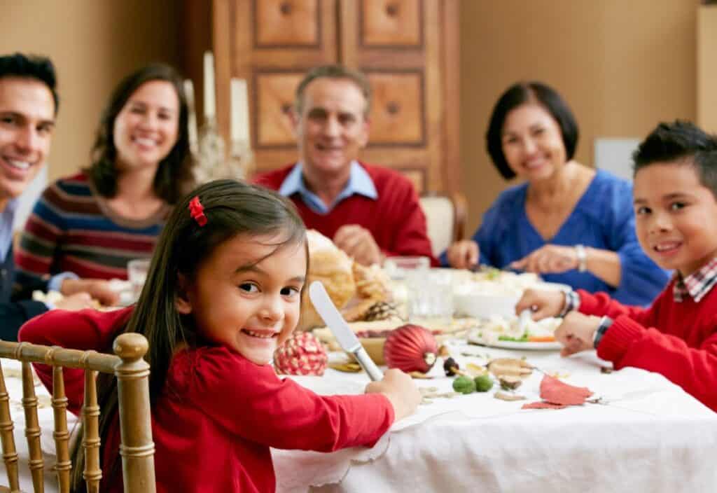 A family of six sits around a dining table, smiling and enjoying a meal together in a warmly lit room.