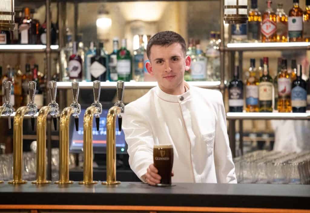 A bartender in a white uniform stands behind a bar, holding a pint of dark beer with a foam head. Bottles of liquor and beer taps are visible in the background.