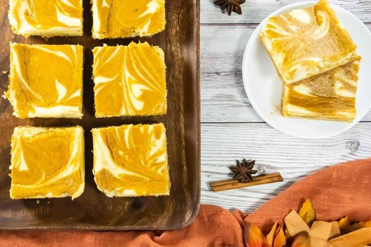 Six pumpkin cheesecake bars with a marbled pattern on a wooden tray, with two bars on a white plate beside cinnamon sticks and star anise on a white wooden table.