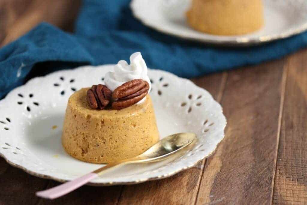 A small round pumpkin custard topped with pecans and whipped cream sits on a white plate with a gold spoon, on a wooden table.