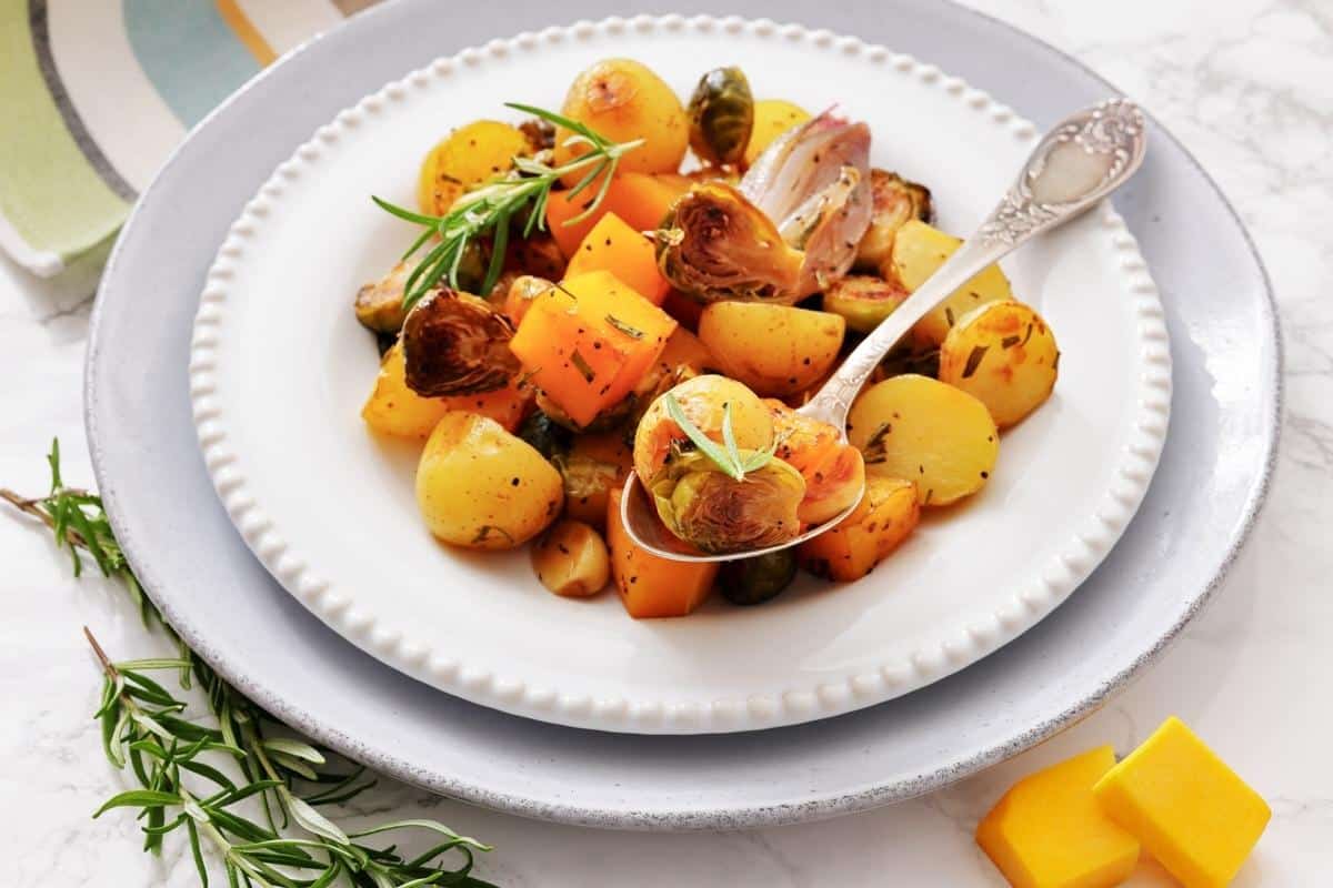 A plate of roasted vegetables including potatoes, Brussels sprouts, and squash, garnished with fresh herbs, with a serving spoon on a white plate.