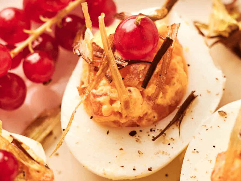 A close-up of a deviled egg topped with crispy garnish and a red currant, with a small bunch of red currants in the background.