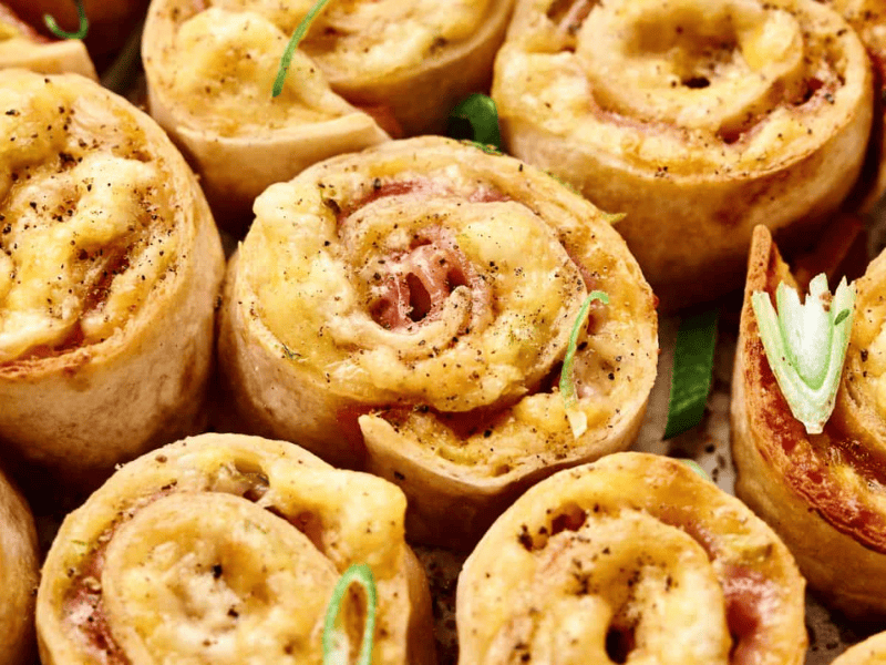 Close-up of baked pinwheel pastries filled with cheese and meat, topped with black pepper and garnished with green herbs.