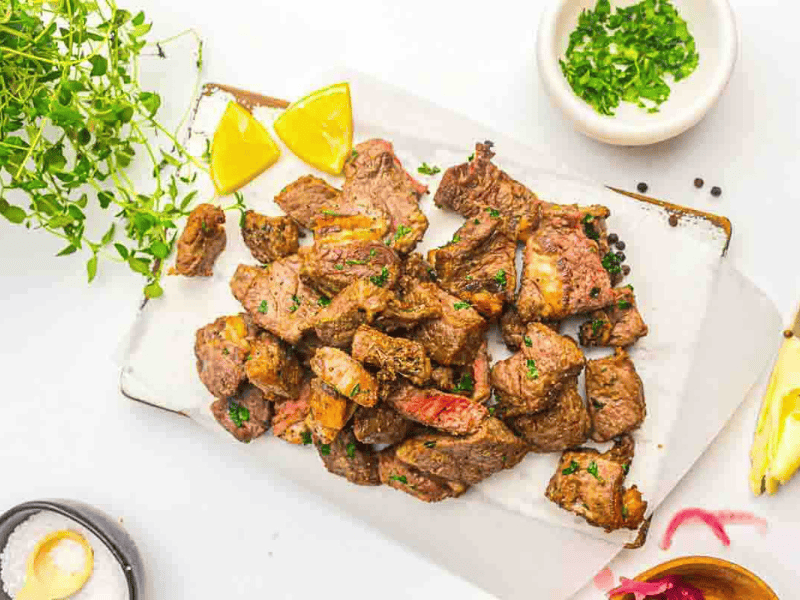 Grilled steak bites garnished with herbs on a white cutting board, served with lemon wedges and surrounded by small bowls of seasoning and chopped herbs.