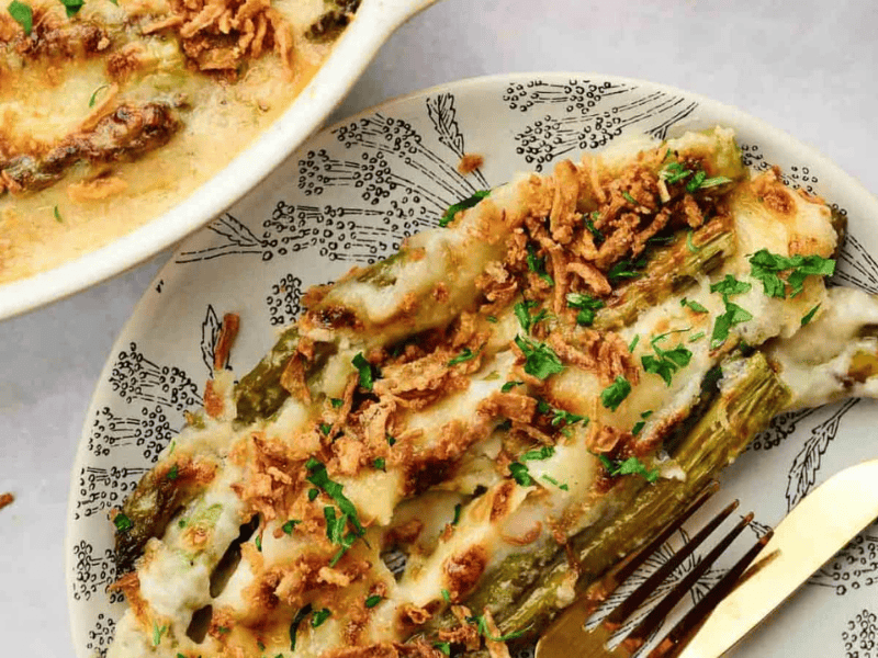 A serving of baked asparagus topped with melted cheese, crispy onions, and chopped herbs, shown on a patterned plate with a gold fork.