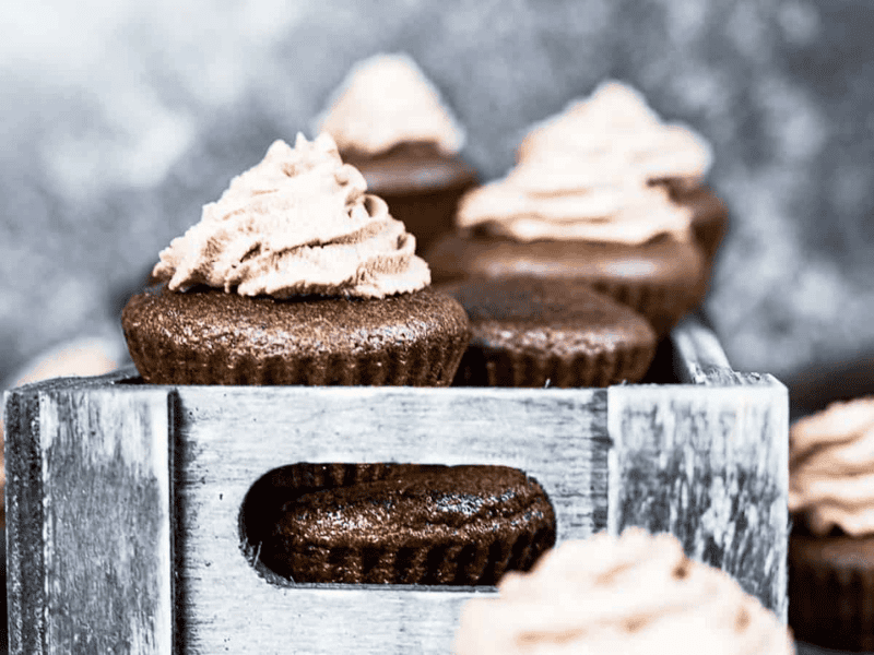 Chocolate cupcakes with swirled light brown frosting are arranged in a rustic wooden box, with a textured gray background.