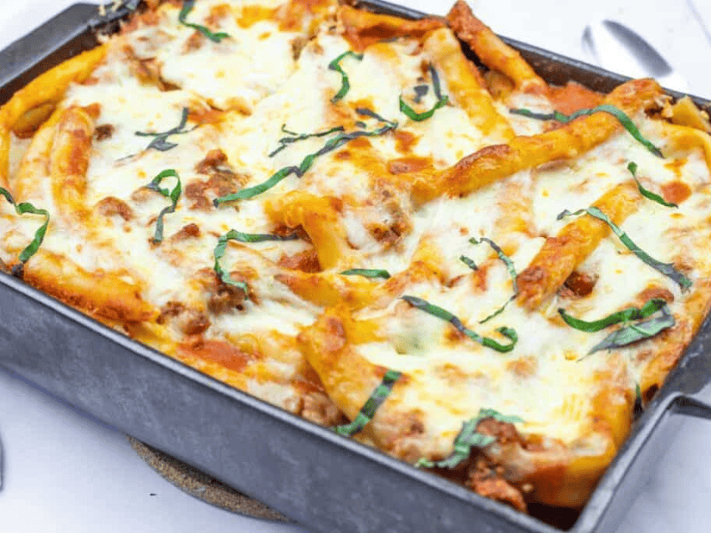 Baked ziti in a rectangular pan topped with melted cheese, tomato sauce, and fresh basil strips.