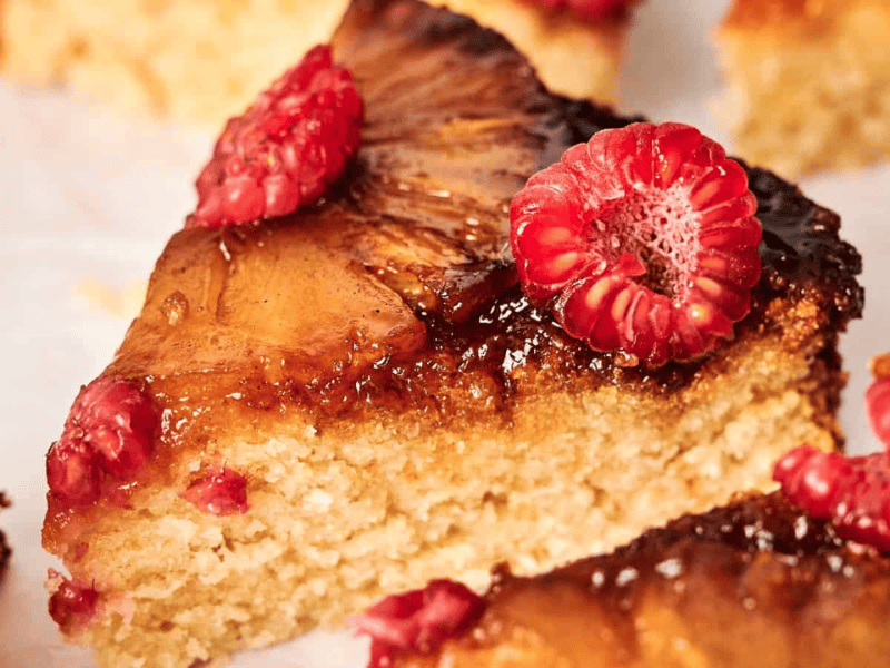 A close-up of a slice of cake topped with caramelized glaze and fresh raspberries.