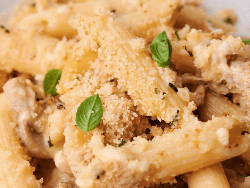 Close-up of baked penne pasta with a creamy sauce, mushrooms, grated cheese, breadcrumbs, and garnished with fresh basil leaves.