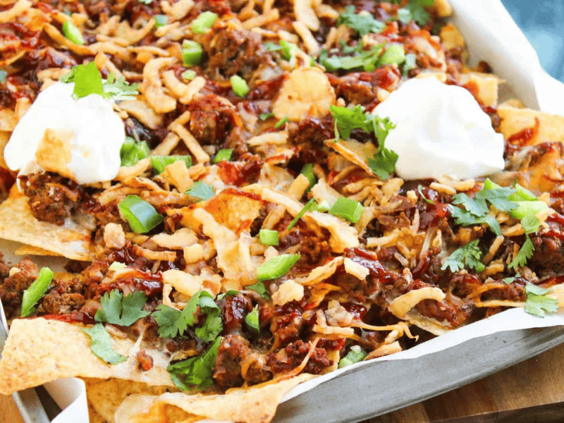 A tray of nachos topped with ground meat, melted cheese, chopped green onions, cilantro, fried onions, barbecue sauce, and two dollops of sour cream.