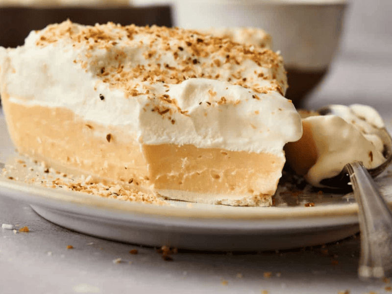 A slice of cream pie with a thick custard filling, topped with whipped cream and toasted coconut flakes, served on a plate.
