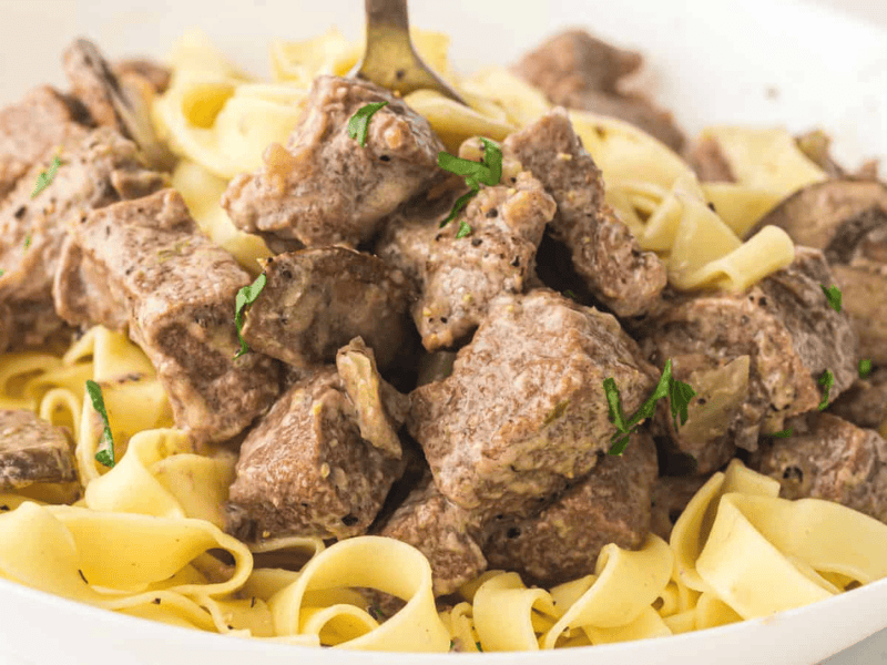 A plate of egg noodles topped with chunks of cooked beef in a creamy sauce, garnished with chopped herbs.