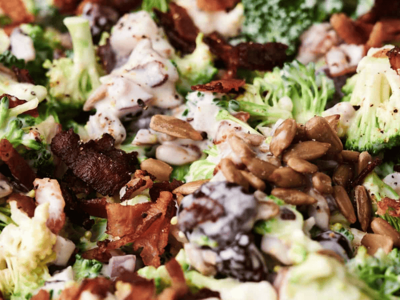Close-up of a broccoli salad topped with sunflower seeds, bacon pieces, and a creamy dressing.