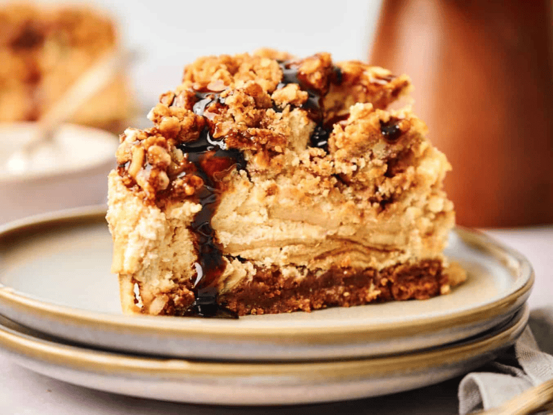 A slice of layered cheesecake with a crumbly topping and a drizzle of dark sauce on a plate.