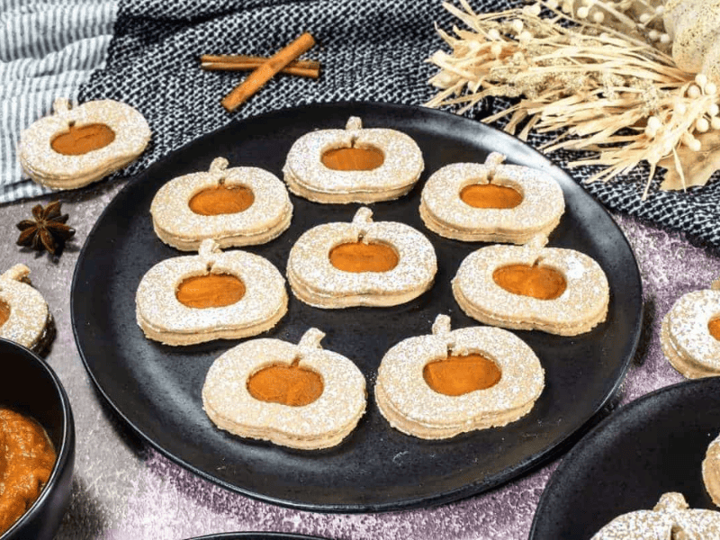 A black plate with twelve pumpkin-shaped sandwich cookies dusted with powdered sugar, each showing a pumpkin-shaped cutout filled with orange jam.