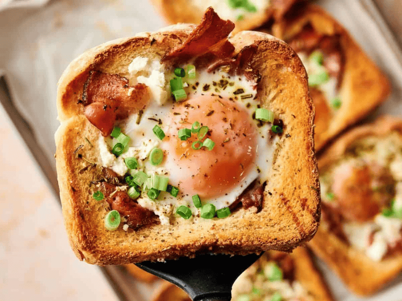 A slice of toasted bread with a cooked egg in the center, topped with bacon, green onions, and herbs, held above a tray of similar pieces.