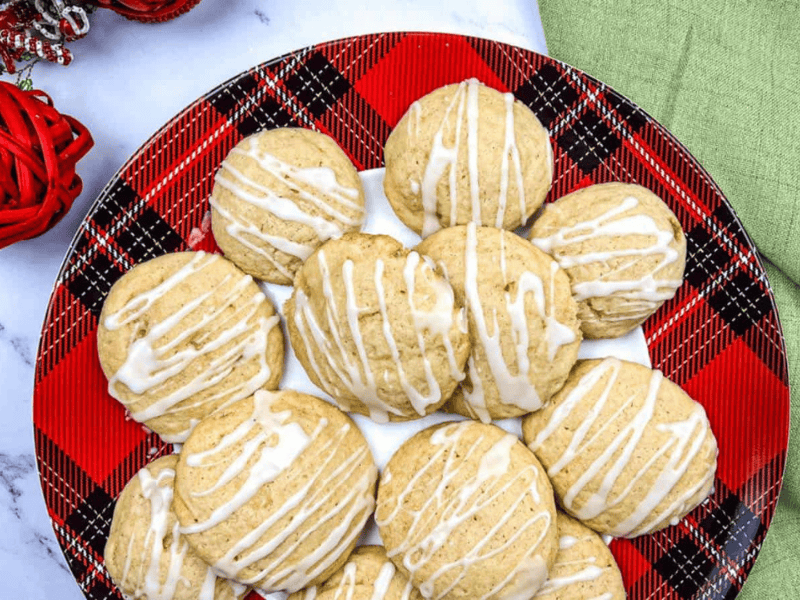 A red plaid plate holds a pile of round, glazed cookies with white icing drizzled on top. A green napkin and red ornaments are visible nearby.
