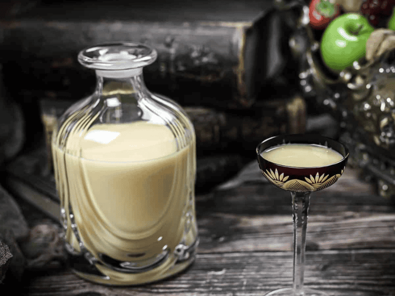 A glass decanter filled with a creamy yellow liqueur sits beside a small, ornate glass with the same drink on a rustic wooden surface.