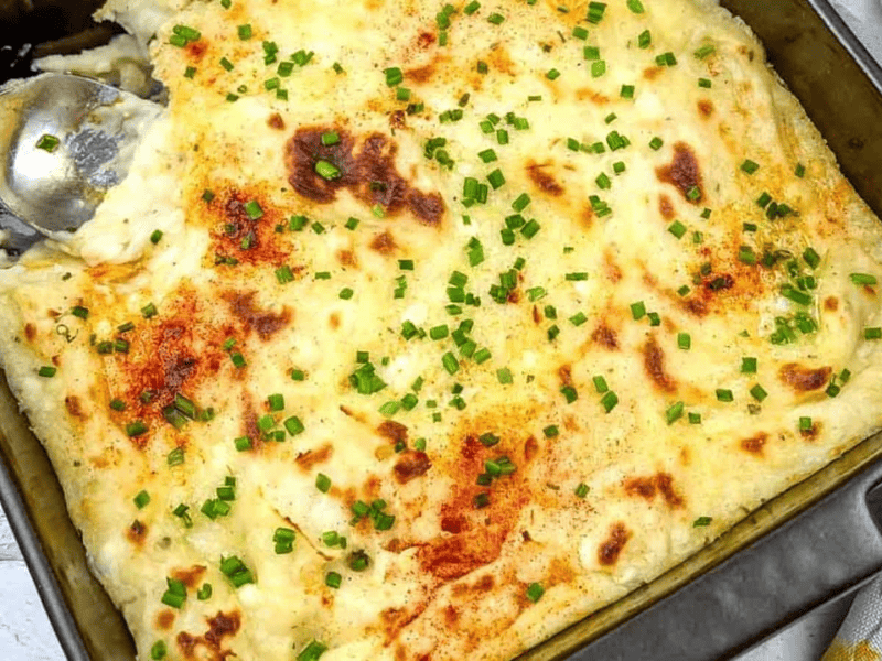 A baked casserole topped with melted cheese, garnished with chopped chives and paprika, with a serving spoon and a portion missing from one corner.