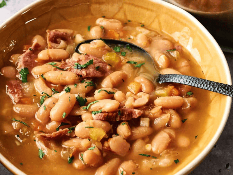 A bowl of white bean soup with pieces of ham, chopped vegetables, and a spoon, garnished with fresh herbs.