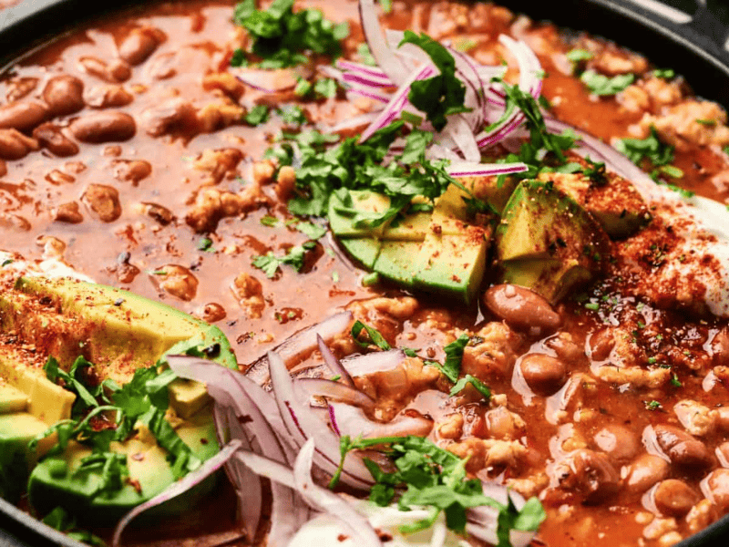 A close-up of a bowl of pinto bean soup topped with sliced avocado, red onion, cilantro, and a sprinkle of chili powder.
