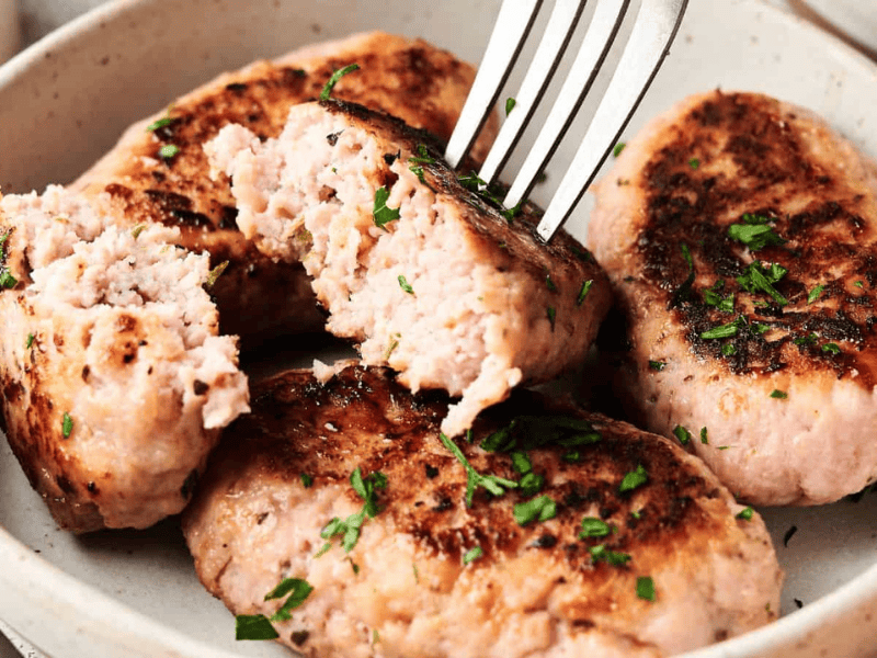 Four browned sausage patties garnished with chopped herbs on a plate, with a fork breaking one open to show the inside.
