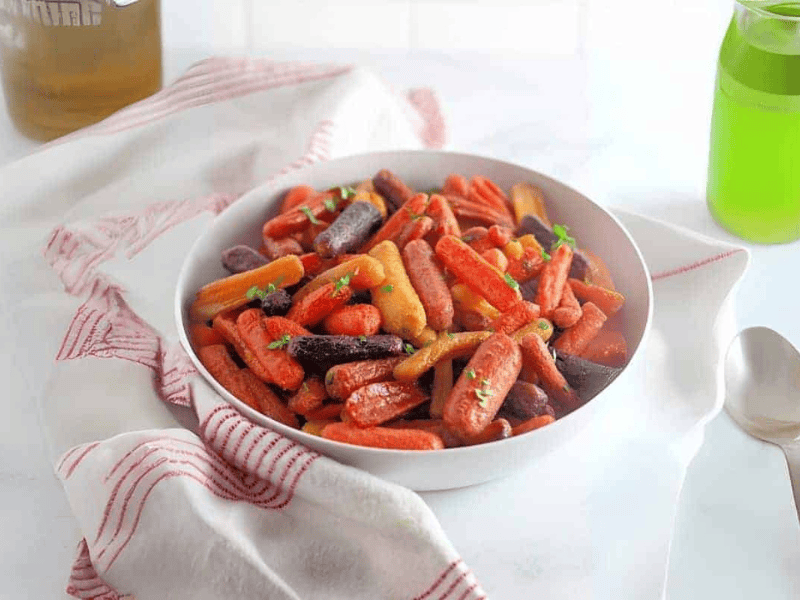A white bowl filled with roasted baby carrots, including orange and purple varieties, garnished with fresh herbs, sits on a white napkin with red stripes.