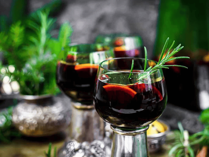 Two glasses of red wine-based drink with orange slices and a sprig of rosemary, placed on a table with greenery and decorative objects in the background.