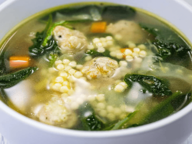 A bowl of clear soup with meatballs, spinach, carrots, and small round pasta in a white bowl.