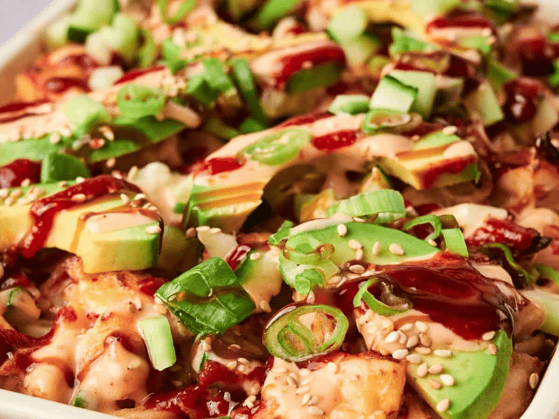Close-up of a dish topped with sliced avocado, green onions, cucumber, sesame seeds, and drizzled with creamy and dark sauces.