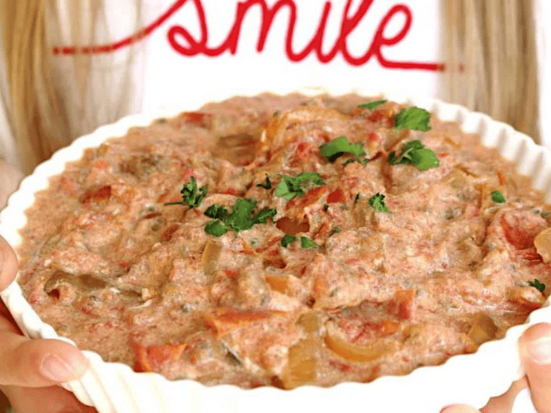 A person holds a white dish filled with creamy, chunky dip or spread, garnished with fresh cilantro; the word 