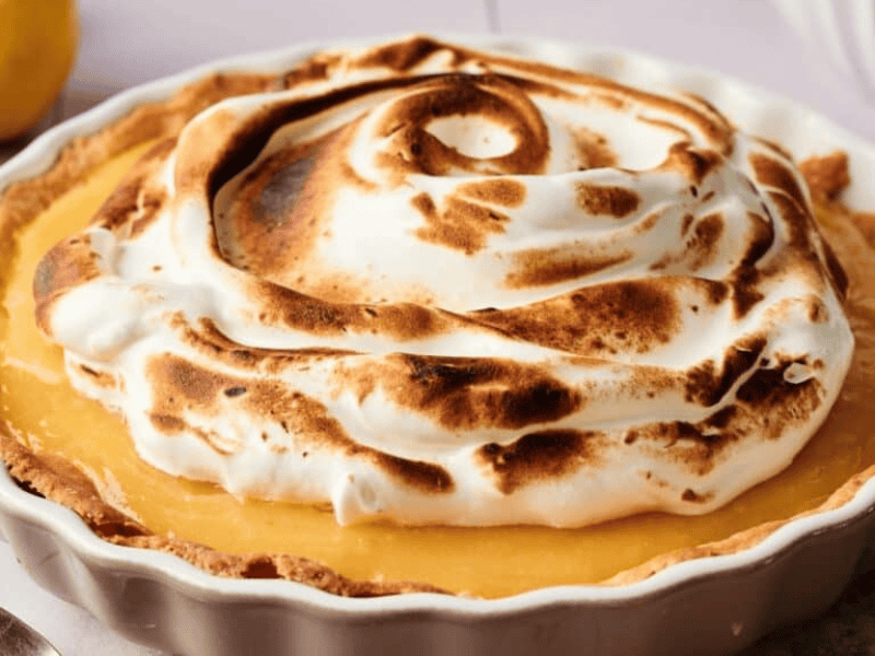 A lemon meringue pie in a white dish, topped with swirled, browned meringue, sits on a light-colored surface next to a lemon.