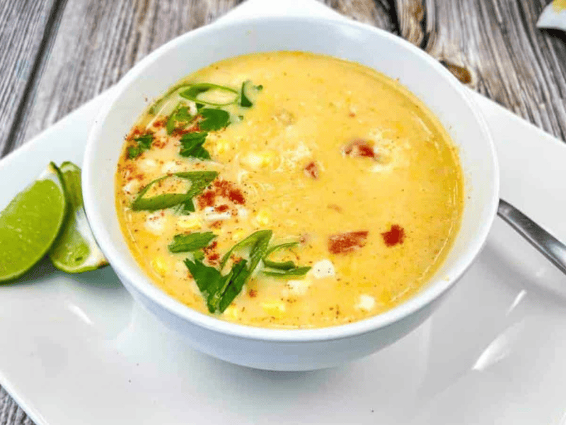 A bowl of creamy soup garnished with chopped green onions, cilantro, and paprika, served with lime wedges on the side.