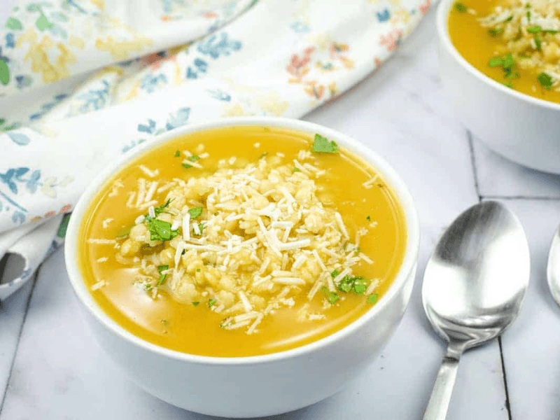 A bowl of Nonna&rsquo;s Pastina Soup topped with shredded cheese and herbs, next to two silver spoons on a marble surface with a floral cloth in the background.