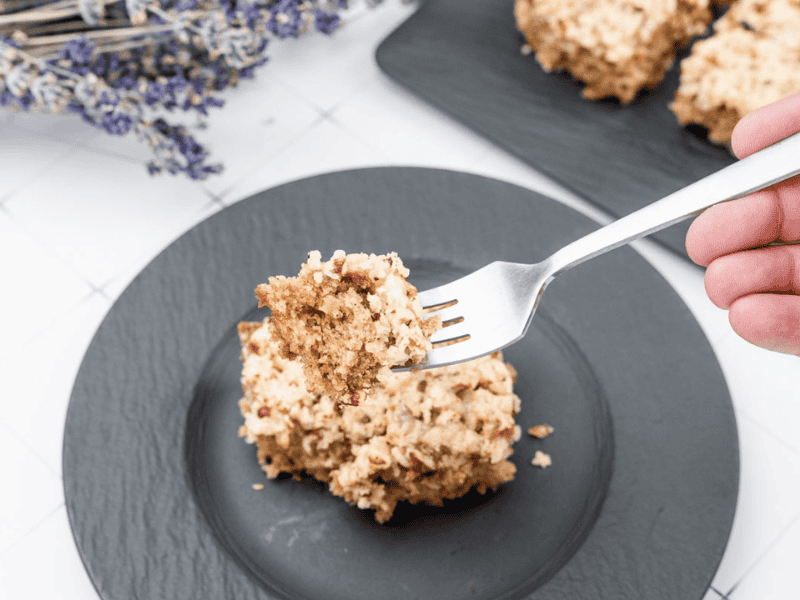 A fork holds a bite of Old Fashioned Lazy Daisy Oatmeal Cake above a black plate with more cake pieces, with dried lavender in the background.