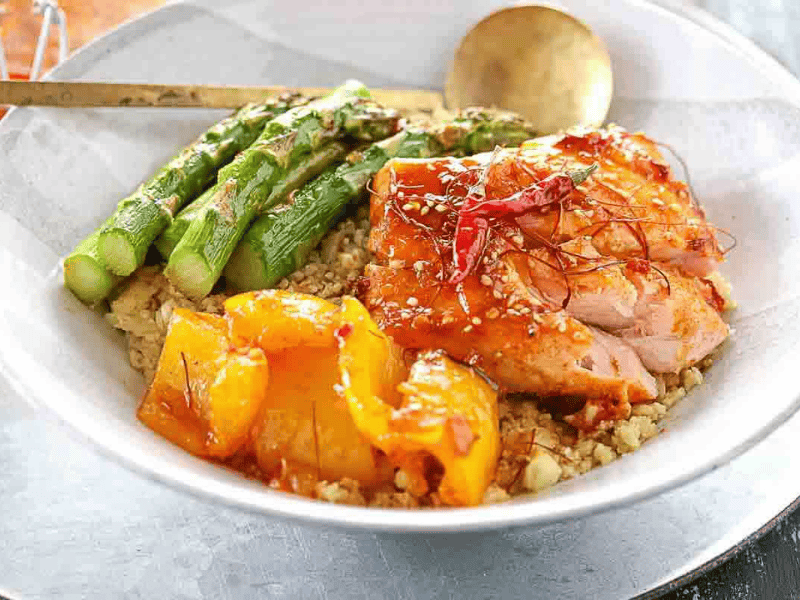 A white bowl with grilled chicken, asparagus, roasted yellow bell peppers, and quinoa, garnished with sesame seeds and chili threads, with a spoon on the side.