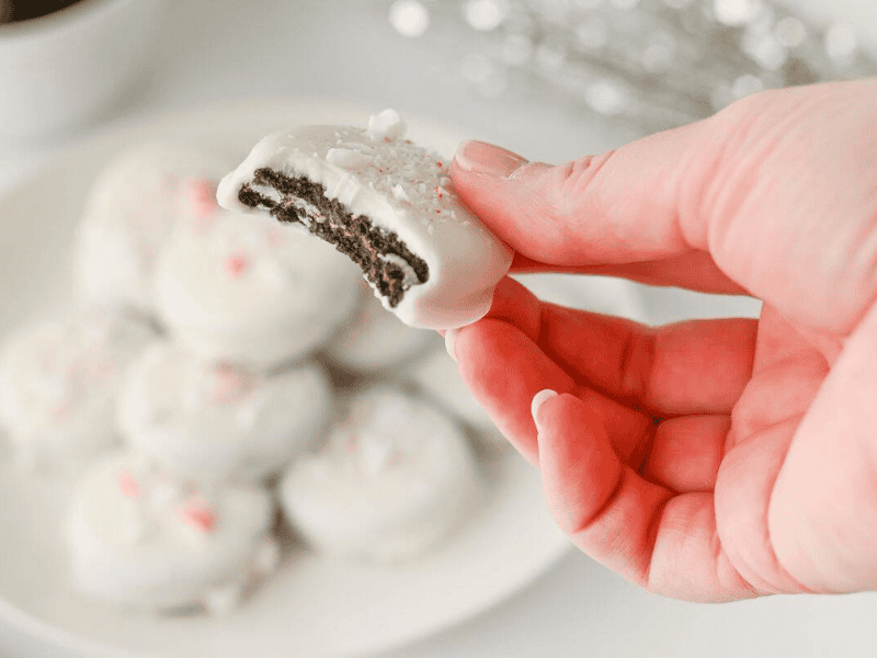 A hand holds a Peppermint Crunch Oreo with a bite taken out, with more cookies on a plate in the background.