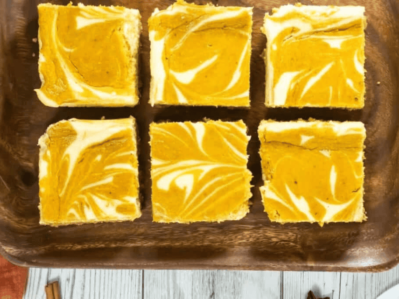Six pumpkin cheesecake bars with a marbled orange and white pattern are arranged on a wooden tray.