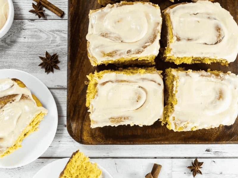Four frosted cinnamon rolls are arranged on a wooden board, with two additional pieces served on white plates beside star anise and cinnamon sticks.